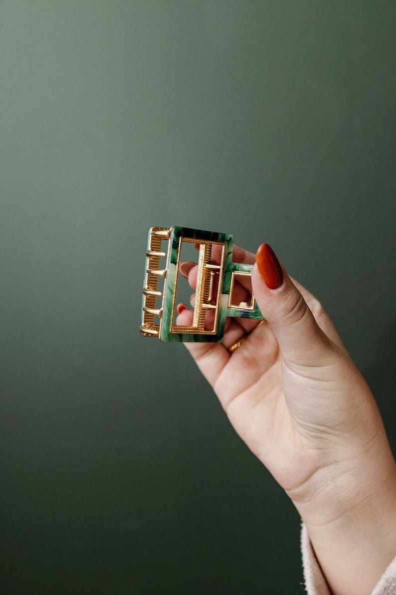 Puede incluir: Una pinza para el pelo de color verde y dorado con forma rectangular. La pinza tiene un patr&oacute;n de m&aacute;rmol y un marco de metal dorado.