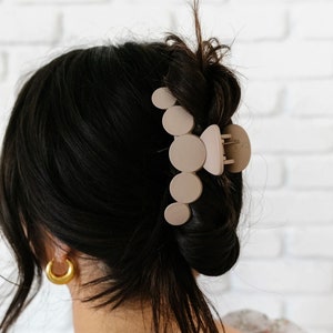 May include: A beige plastic hair claw with a rounded, abstract design. The claw is holding a woman's dark brown hair in a bun.