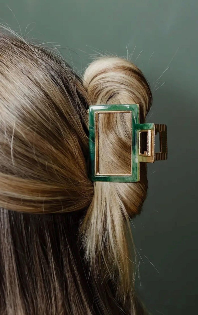 Puede incluir: Una pinza para el pelo de color verde y dorado con un dise&ntilde;o rectangular. La pinza est&aacute; hecha de pl&aacute;stico y tiene un marco de metal dorado.