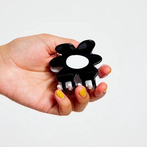May include: Black and white flower-shaped hair clip with a white centre. The clip has five prongs on the bottom.