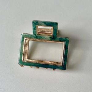 Puede incluir: Una pinza para el pelo verde y dorada con un marco rectangular. La pinza tiene un patr&oacute;n de m&aacute;rmol y un borde dorado.