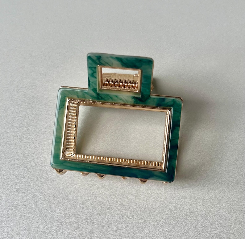 Puede incluir: Una pinza para el pelo verde y dorada con un marco rectangular. La pinza tiene un patr&oacute;n de m&aacute;rmol y un borde dorado.
