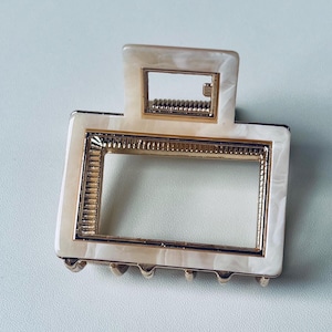 Puede incluir: Una pinza para el pelo blanca y dorada con un marco rectangular. La pinza tiene un patr&oacute;n de m&aacute;rmol y un borde dorado.