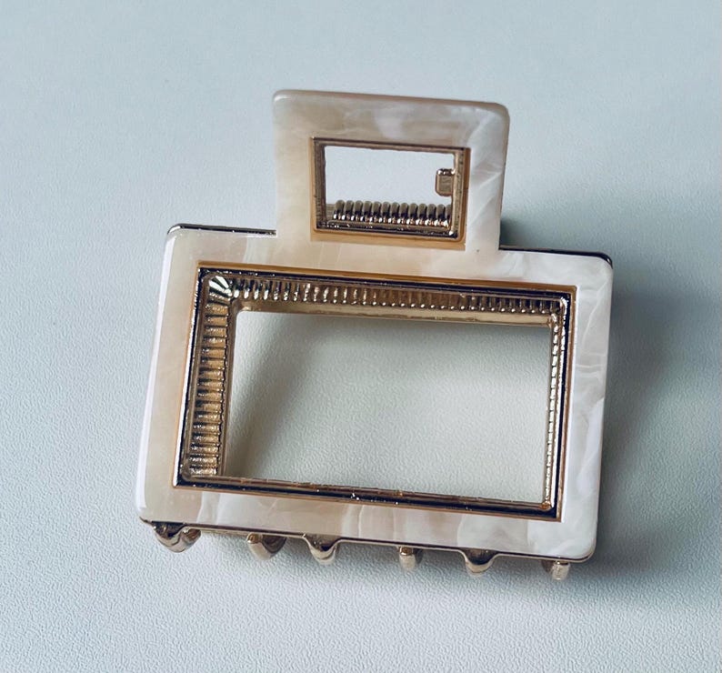 Puede incluir: Una pinza para el pelo blanca y dorada con un marco rectangular. La pinza tiene un patr&oacute;n de m&aacute;rmol y un borde dorado.