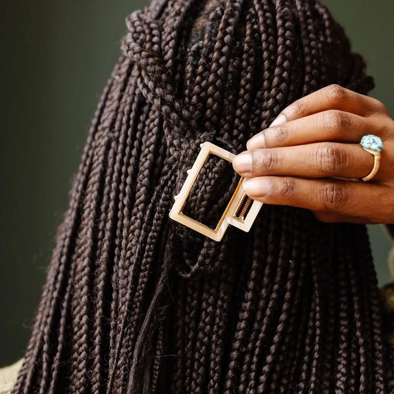 Puede incluir: Una pinza para el pelo de color dorado con forma rectangular. La pinza se utiliza para sujetar una secci&oacute;n de cabello largo, trenzado de color marr&oacute;n oscuro.