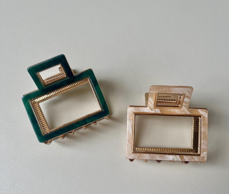 Puede incluir: Dos pinzas para el pelo, una verde y otra beige, con ribete dorado. Ambas pinzas tienen forma rectangular con una abertura rectangular m&aacute;s peque&ntilde;a en el centro.