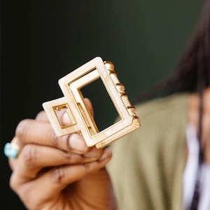 Puede incluir: Una pinza para el pelo de color dorado y blanco con forma rectangular y una abertura cuadrada m&aacute;s peque&ntilde;a en el centro.