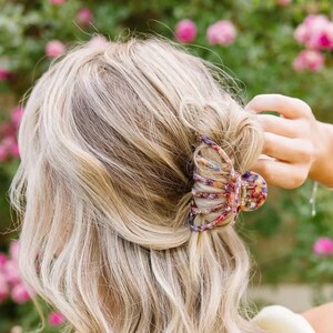 May include: A large, colourful hair claw with a floral pattern. The claw is made of plastic and has a claw-like shape. It is being used to hold a woman's blonde hair in a half-up hairstyle.