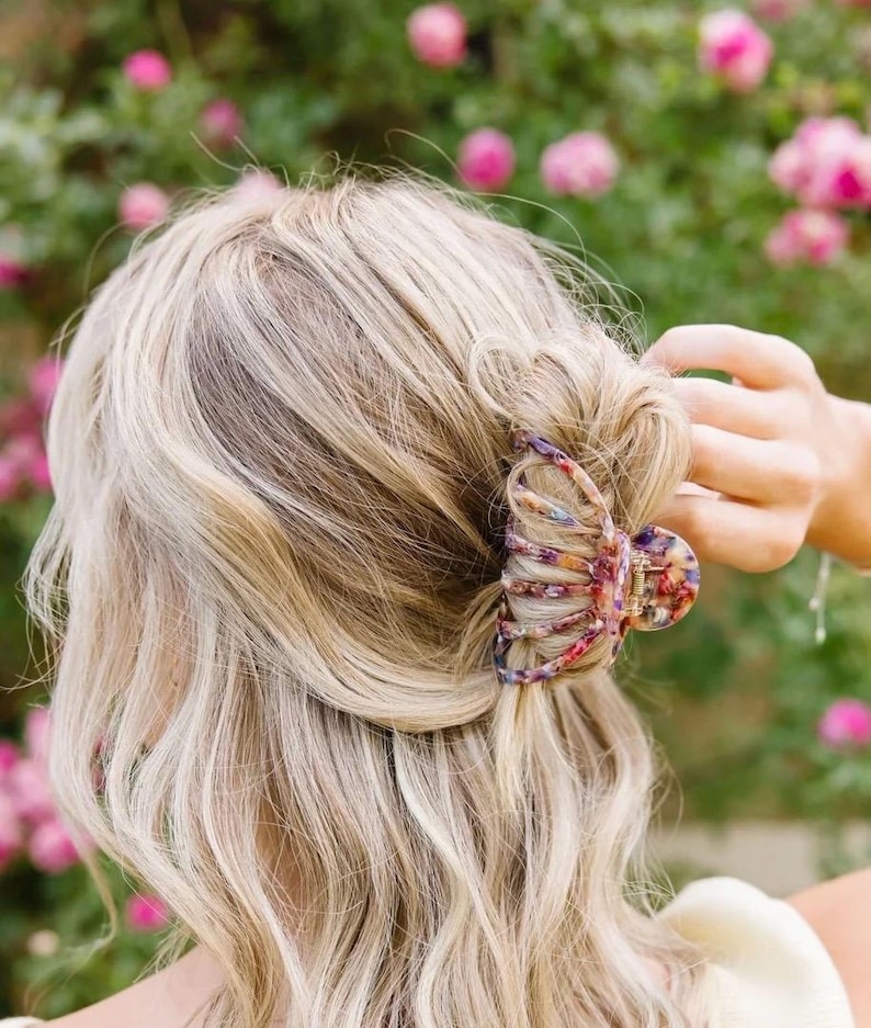 May include: A large, colourful hair claw with a floral pattern. The claw is made of plastic and has a claw-like shape. It is being used to hold a woman's blonde hair in a half-up hairstyle.