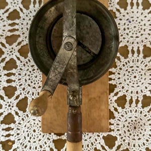 May include: An antique, hand-cranked kitchen tool, likely a butter churn or similar device. It features a circular metal component with a handle and wooden grip, mounted on a wooden base. The metal has a weathered, aged appearance.