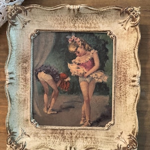 May include: A framed vintage print depicts two young ballerinas in a soft, muted color palette. The frame is ornate, with a distressed white finish and gold accents. The artwork shows the ballerinas in pink and purple tutus.