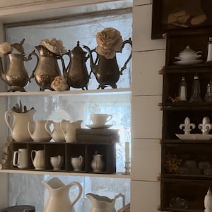 May include: A white shelf unit with a variety of white ceramic pitchers and teapots, along with silver teapots decorated with silk flowers. The shelf is against a white wall with a window behind it.