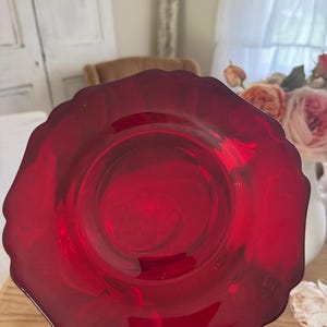 May include: A translucent ruby red glass plate featuring a smooth, circular center and a decorative scalloped edge. The plate is crafted from deep red glass that allows light to pass through, highlighting its vibrant color and glossy finish.