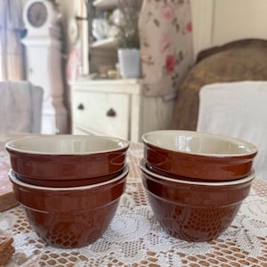 May include: A stack of four brown ceramic bowls with a cream-colored interior. The bowls have a classic, rounded shape and are arranged on a white lace tablecloth. The bowls are likely for serving food.