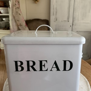 May include: A white metal bread box with a square shape and a removable lid featuring a curved handle. The front of the container displays the word BREAD in bold, black, uppercase lettering. The finish is smooth and glossy with clean edges.