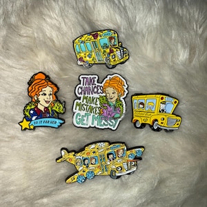 May include: Set of five enamel pins featuring characters from the animated television series The Magic School Bus. The pins depict the bus in various forms, including a regular school bus, a flying bus, and a bus with a message that reads "Take chances, make mistakes, get messy".