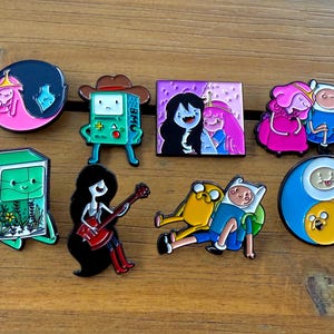 May include: A collection of enamel pins featuring characters from the animated series Adventure Time. The pins showcase characters like Princess Bubblegum, BMO, Marceline, Finn, and Jake in various poses and designs. The pins are colorful and have a glossy finish.