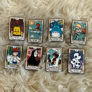 May include: A collection of eight rectangular enamel pins featuring anime-style illustrations. Each pin has a gold border and depicts a tarot card design with titles like "The Devil," "The Star," and "The Fool." The pins are set against a fluffy, white background.