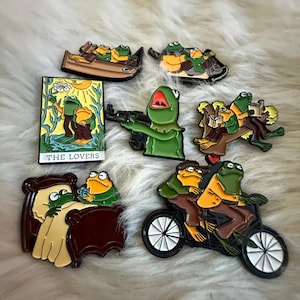 May include: Set of 7 enamel pins featuring green frogs in various scenes. The pins include a frog holding a gun, a frog in a boat, a frog in bed, a frog riding a bike, a frog reading a book, and a frog in a tarot card depicting "The Lovers".