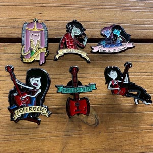 May include: A collection of enamel pins featuring characters and quotes from a cartoon series. The pins include characters playing guitars, and phrases like "You Rock!" and "Everything Stays". The pins are in various colors, including red, black, and pink.