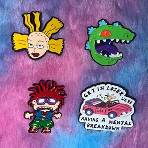 May include: Four colorful enamel pins featuring characters from a cartoon series. Includes a blonde girl, a green dinosaur with an open mouth, a red-haired boy, and a pink car with text that reads "Get in loser we're having a mental breakdown."