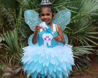 May include: A young girl dressed as a tooth fairy, wearing a blue and white tutu with silver glitter, a crown, and fairy wings. She is holding a wand with a tooth on the end.