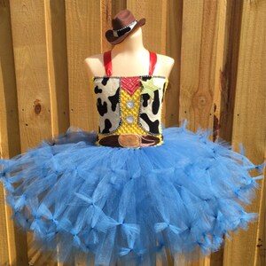 May include: A blue tulle tutu with a yellow and brown cowboy vest and a brown cowboy hat. The vest has a white and black cow print and a red bandana. The costume is perfect for a child's dress-up party.