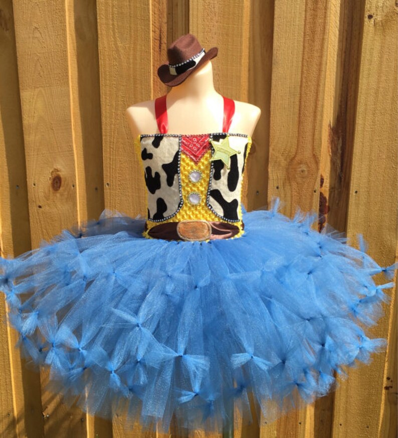 May include: A blue tulle tutu with a yellow and brown cowboy vest and a brown cowboy hat. The vest has a white and black cow print and a red bandana. The costume is perfect for a child's dress-up party.