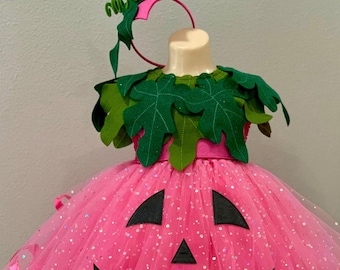 May include: Pink tulle pumpkin costume with a black jack-o'-lantern face and green felt leaves. The costume includes a pink headband with green felt leaves and a brown stem.