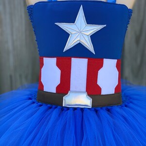 May include: A blue tulle tutu with a red, white, and blue Captain America costume top. The top features a silver star and a brown belt with a silver buckle.