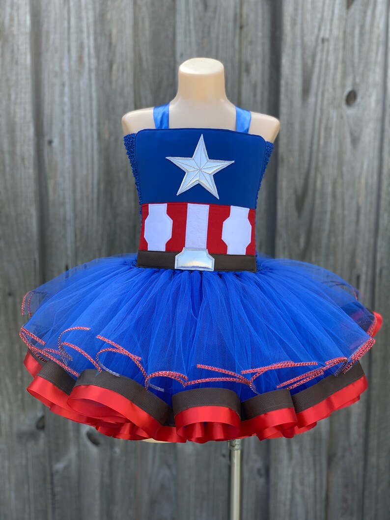 May include: A blue tulle tutu with red and brown ribbon trim. The top of the tutu is a blue and red fabric with a white star and a brown belt.