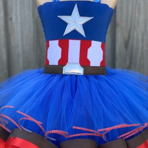 May include: A blue tulle tutu with red and brown ribbon trim. The top of the tutu is a blue and red fabric with a white star and a brown belt.