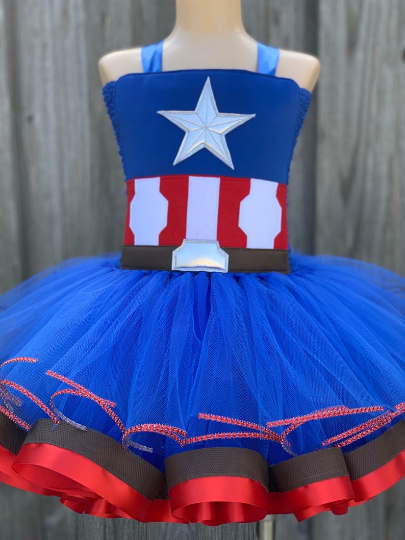 May include: A blue tulle tutu with red and brown ribbon trim. The top of the tutu is a blue and red fabric with a white star and a brown belt.