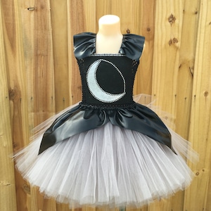 May include: A black and silver tulle tutu dress with a crescent moon design on the bodice. The bodice is made of black satin with a silver glitter crescent moon. The skirt is made of layers of tulle.