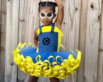 May include: A young girl dressed as a minion for Halloween. She is wearing a blue tulle tutu with yellow ribbon trim, a denim overall dress with a yellow top and a minion logo, and silver goggles.