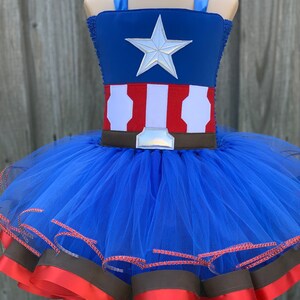 May include: A blue tulle tutu with red and brown ribbon trim. The top of the tutu is a blue and red Captain America costume with a silver star.