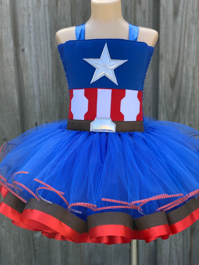 May include: A blue tulle tutu with red and brown ribbon trim. The top of the tutu is a blue and red Captain America costume with a silver star.