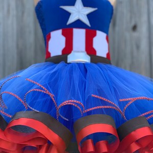 May include: A blue tulle tutu with red and brown ribbon trim. The top of the tutu is a blue and red Captain America costume with a white star.