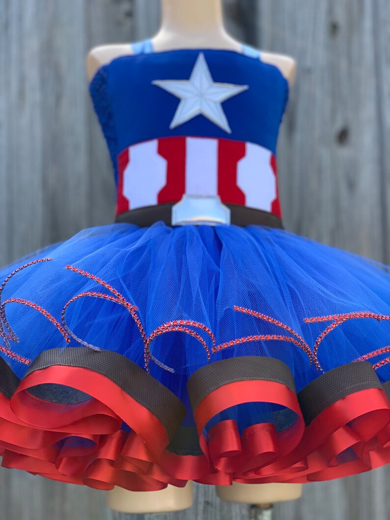 May include: A blue tulle tutu with red and brown ribbon trim. The top of the tutu is a blue and red Captain America costume with a white star.