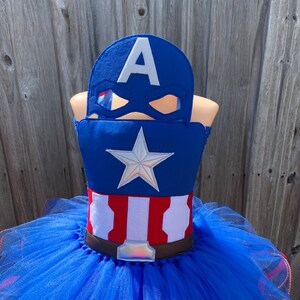 May include: A blue tulle tutu with a red, white, and blue Captain America costume top. The costume top features a white star and the letter "A" on a blue background. A brown belt with a silver buckle is included.