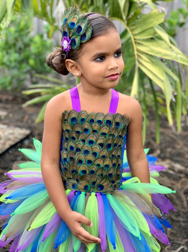 May include: A young girl wearing a peacock-themed tutu dress with a green, blue, and purple tulle skirt. She is wearing a peacock feather headband with a purple flower accent.