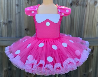 May include: A pink tulle tutu with white polka dots and a white lace trim. The tutu has a pink bodice with white polka dots and a white collar with a single button.