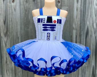 Può includere: Tutu in tulle bianco con bordino di nastro blu e un'applicazione di personaggio R2-D2 bianco e blu sul corpetto.