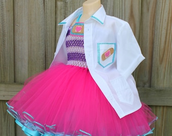 May include: A pink tulle tutu with a blue ribbon trim and a white button-up shirt with a patch pocket featuring a bandage and a heart. The shirt has a blue trim around the collar and cuffs.