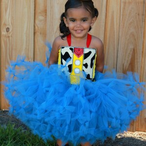 May include: A young girl wearing a blue tulle tutu and a cowboy hat with a star. She is wearing a red and white cow print top with a yellow sheriff's star and a red bandana.