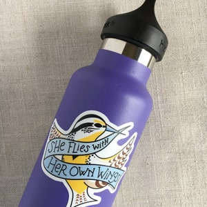 A single She Flies with Her Own Wings vinyl sticker on a purple water bottle.