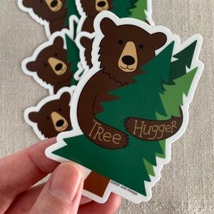 May include: A brown bear hugging a green pine tree with the text "Tree Hugger" on the tree. The bear is smiling and has a brown nose. The tree has a brown trunk and green needles.