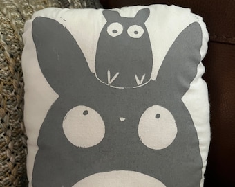 May include: A rectangular pillow featuring a gray cartoon character design on a white background. The design includes a large, round-eyed creature with a smaller creature perched on top. The pillow is resting on a textured, neutral-toned blanket.