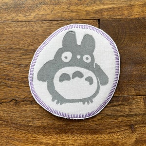 May include: A white fabric patch with a gray embroidered image of a cartoon character with large eyes and ears. The patch has a purple stitched border.