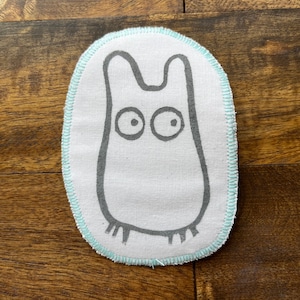 May include: A white fabric patch with a gray outline of a cartoon creature with large eyes. The patch has a light blue stitched border and is oval shaped.